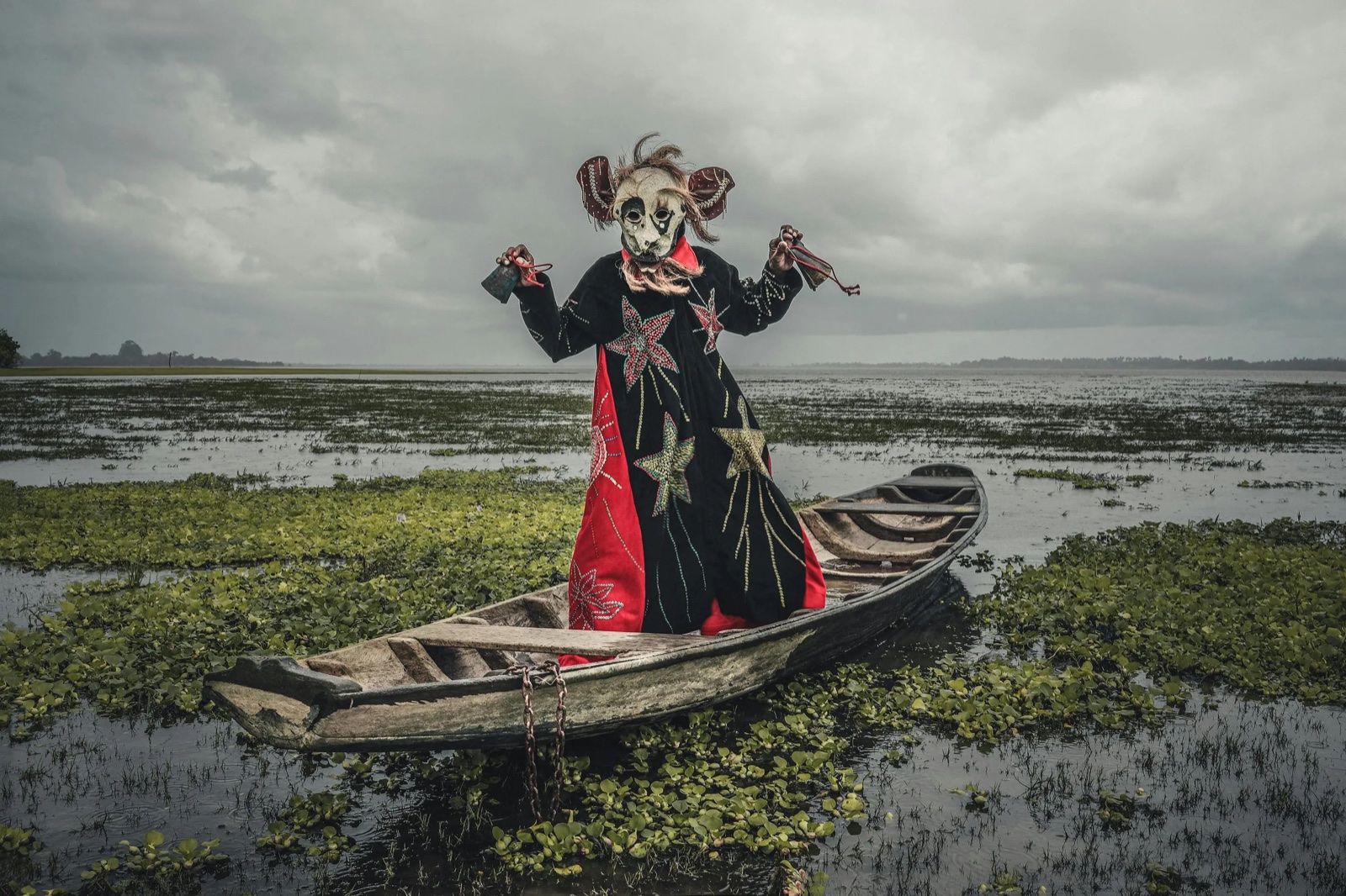 Márcio Vasconcelos — photographic work of a masked figure standing in a canoe beneath a storm-darkened sky by Márcio Vasconcelos